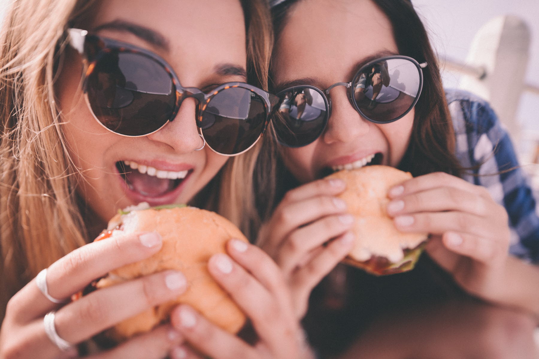 Två  unga kvinnor med långt hår och solglasögon äter hamburgare utomhus