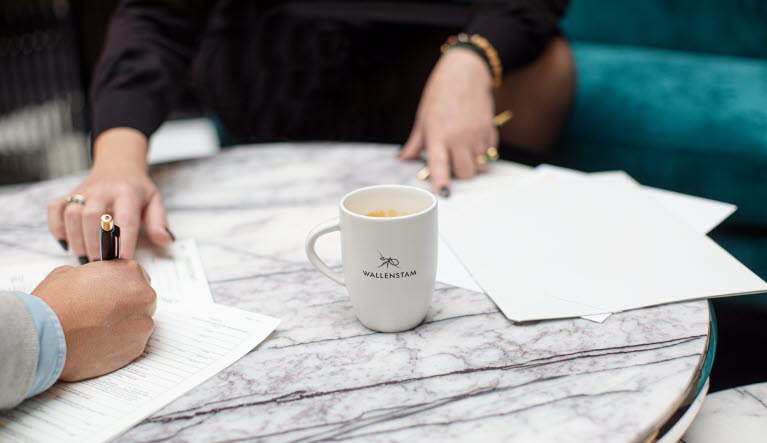 Blanketter och en kaffekopp med Wallenstamloggan på ett bort. Händer som håller i en penna och skriver på blanketterna.
