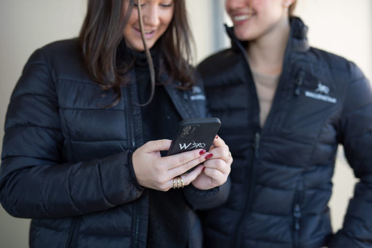 Två kvinnor står utomhus den ena scrollar på en mobiltelefon med Wallenstams logga på skalet.