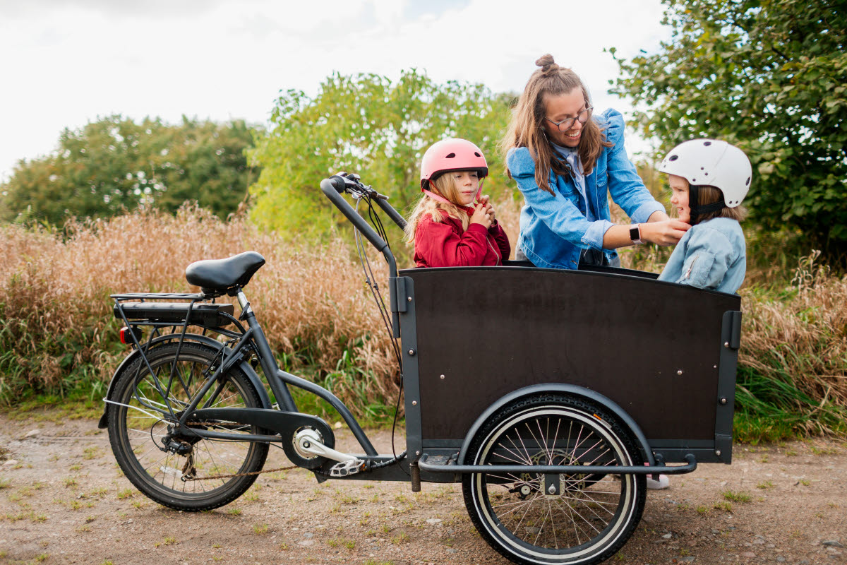Närbild på en mamma som hjälper sitt barn att knäppa sin cykelhjälm. Det är två barn som sitter i en lastcykel utomhus med skog i bakgrunden.