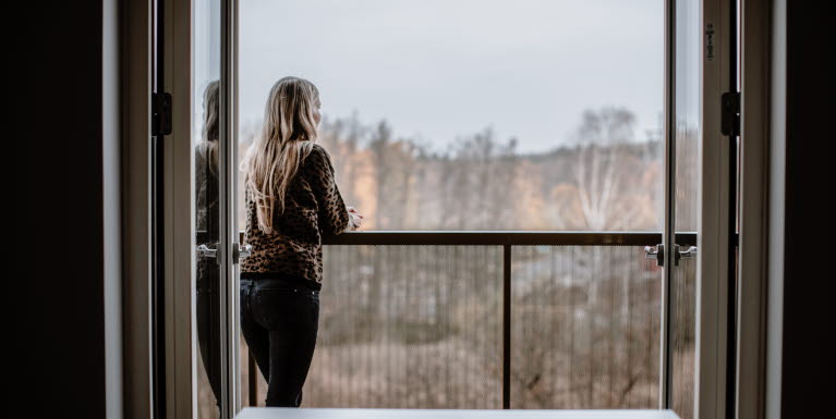 En kvinna med långt hår står på en balkong och tittar ut över ett bostadsområde.
