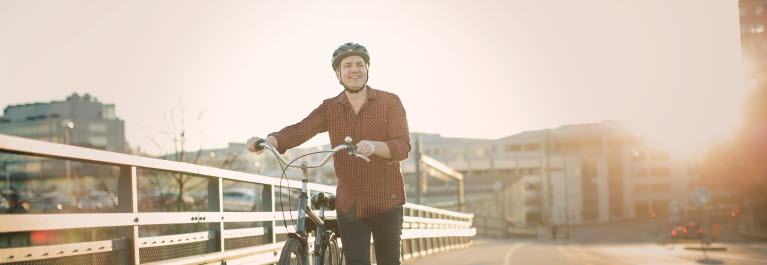 En leende man leder en cykel över en bro. Mannen bär cykelhjälm , jeans och en en röd och svartrutig skjorta