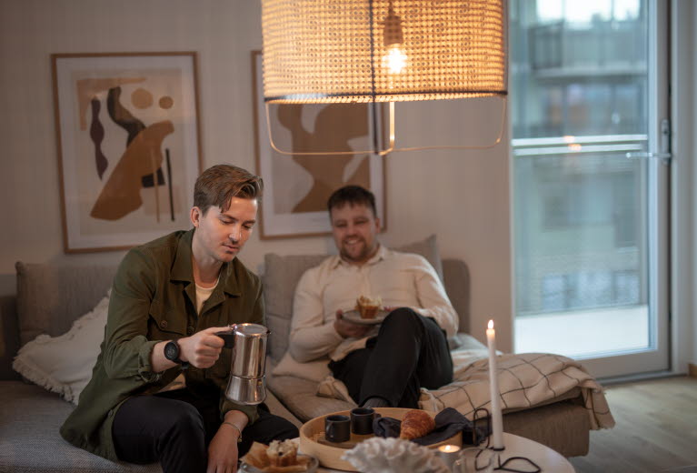 Två yngre killar sitter i soffan i ett vardagsrum i en lägenhet. De pratar, dricker kaffe och äter muffins.