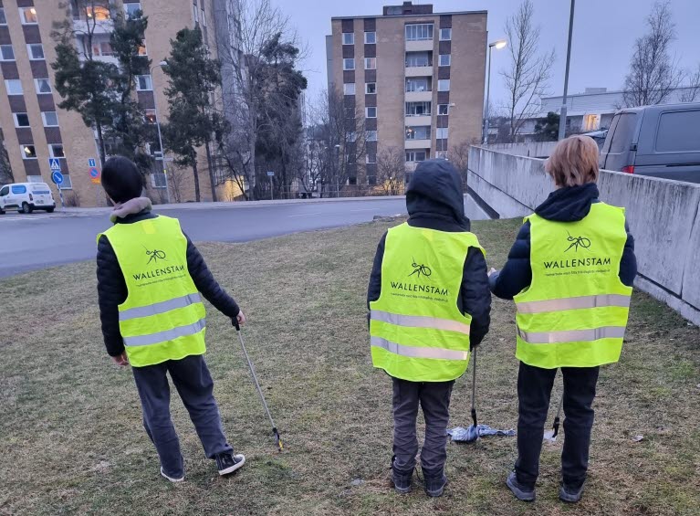 Tre ungdomars om plockar skräp på en gräsmatta. Ungdomarna har på sig gula västar med Wallenstams logotyp