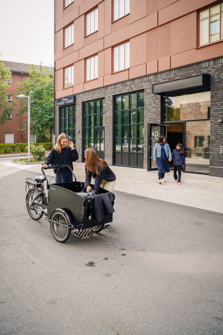 Lastcykel med två kvinnor framför en fastighet samt mor och dotter i bakgrunden